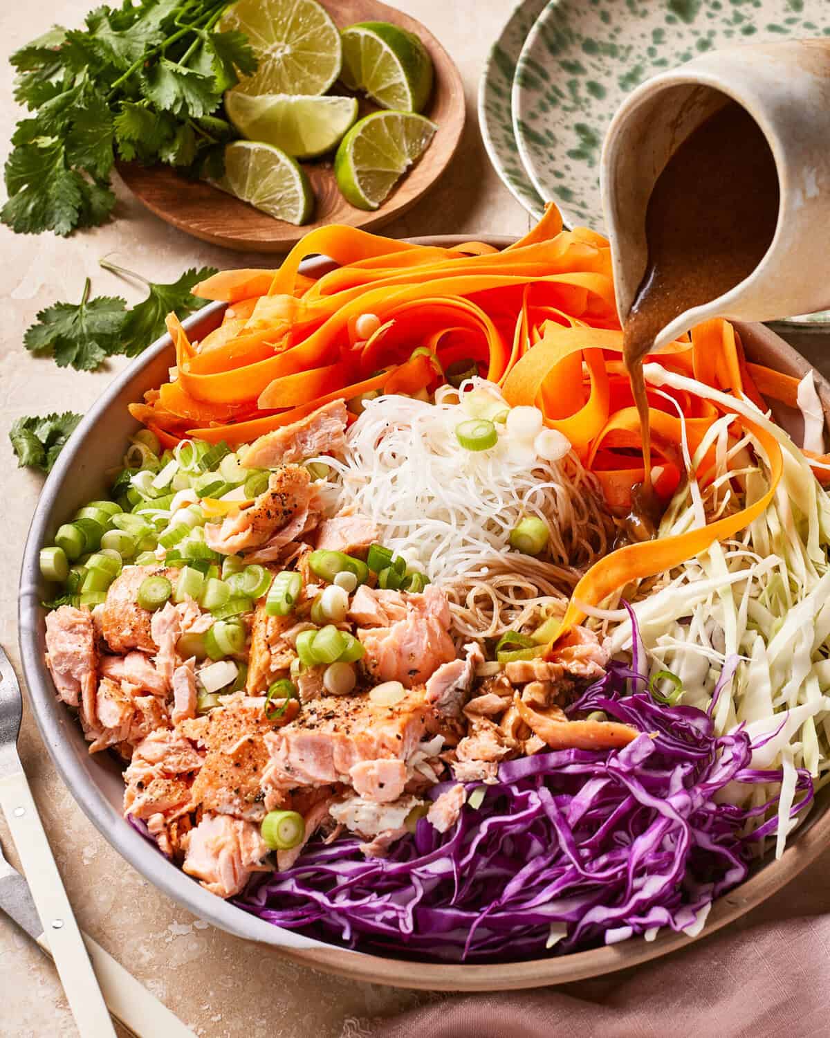 A bowl filled with rice noodles, shredded carrots, purple cabbage, white cabbage, sliced green onions, and cooked salmon chunks, with dressing being poured on top. Fresh lime wedges and cilantro are on the side.