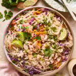 A bowl of salmon crunch salad with shredded purple cabbage, carrots, cooked salmon, cilantro, and lime wedges, garnished with green onions, sits on a table next to a plate of lime halves and fresh cilantro.