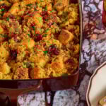 A casserole dish filled with golden-brown cornbread stuffing garnished with herbs and cranberries, sitting on a marbled surface next to an empty plate and utensils.