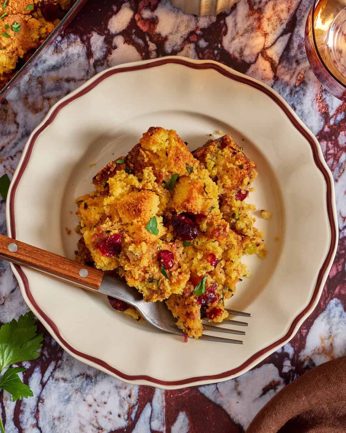 A plate of cornbread stuffing mixed with cranberries and herbs sits on a decorative plate with a fork, placed on a marble surface next to a sprig of parsley.