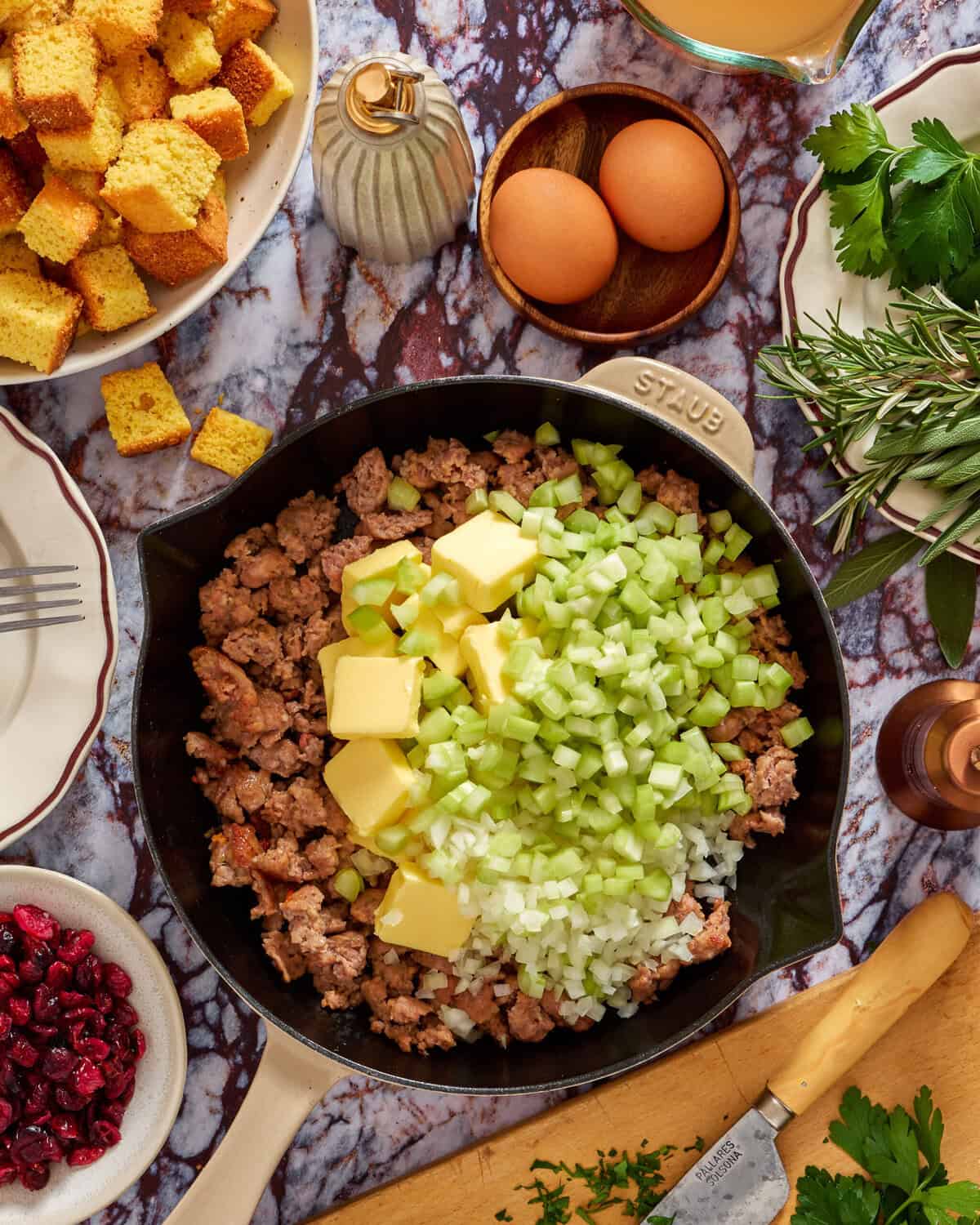 A skillet filled with cooked sausage, topped with cubes of butter, chopped celery, and onions sits on a marble countertop, surrounded by cornbread cubes, eggs, herbs, cranberries, and a knife with chopped parsley.