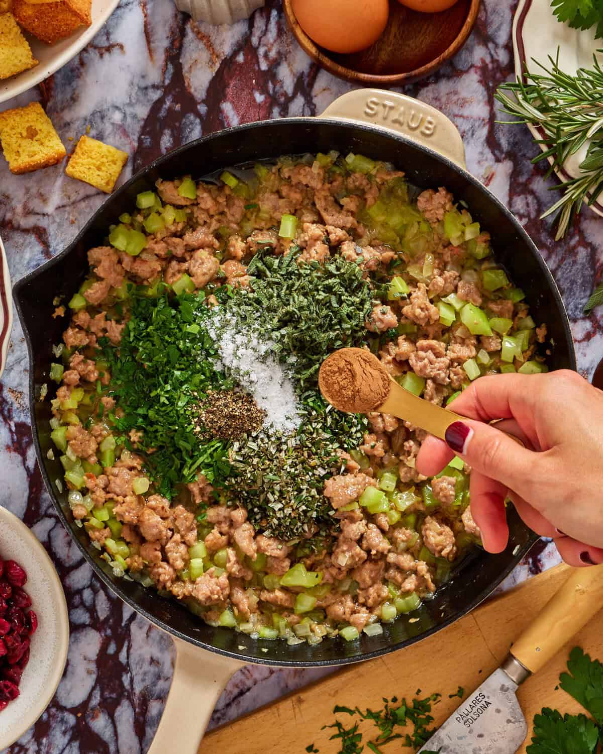 A hand sprinkles ground spices from a measuring spoon into a skillet with cooked sausage, chopped celery, and fresh herbs. Other ingredients, cornbread, eggs, cranberries, and fresh herbs, are visible around the skillet.