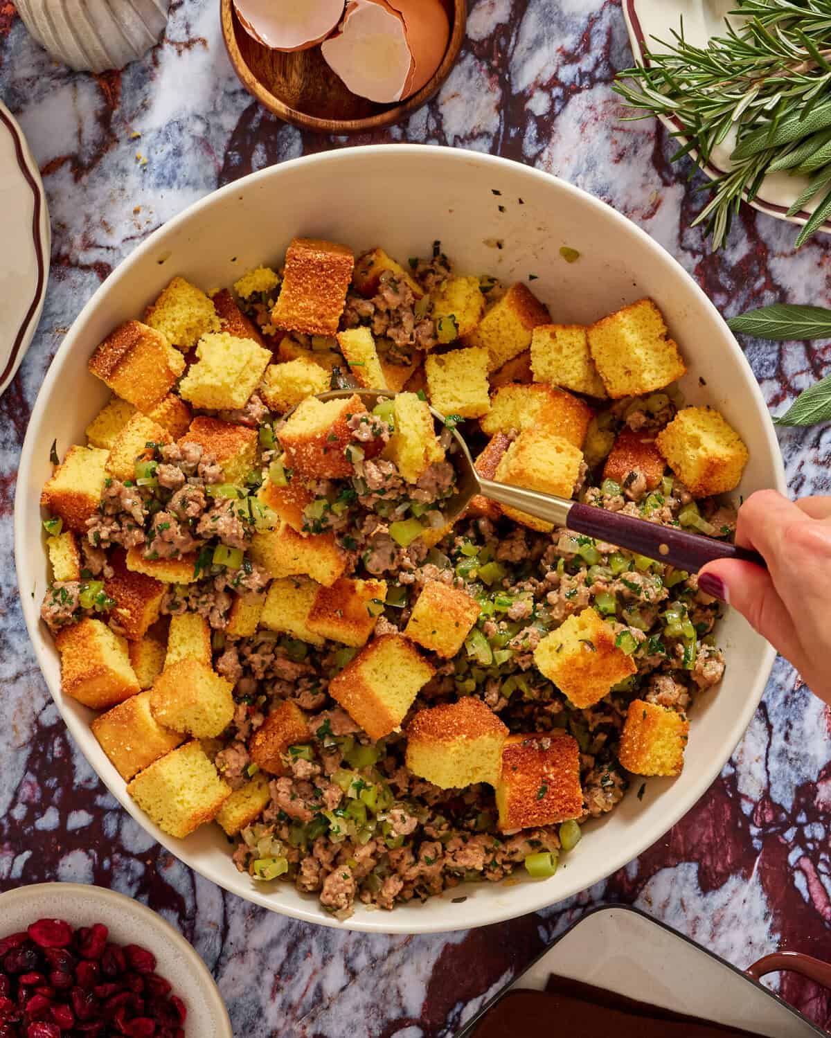 A large white bowl filled with cornbread stuffing, featuring golden cornbread cubes, sausage, celery, and herbs. A hand with a spoon is mixing the stuffing. Fresh herbs and eggshells are visible nearby on a marble countertop.