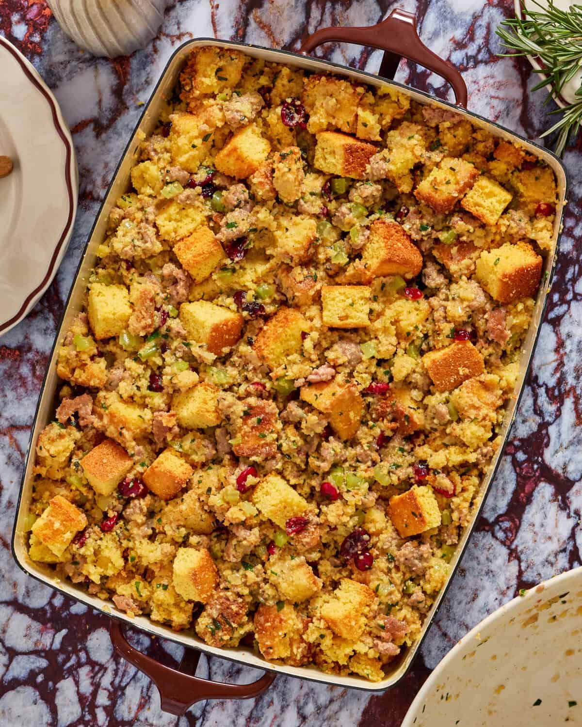 A casserole dish filled with golden-brown cornbread stuffing mixed with sausage, celery, and cranberries on a marble countertop, with a plate and herbs nearby.