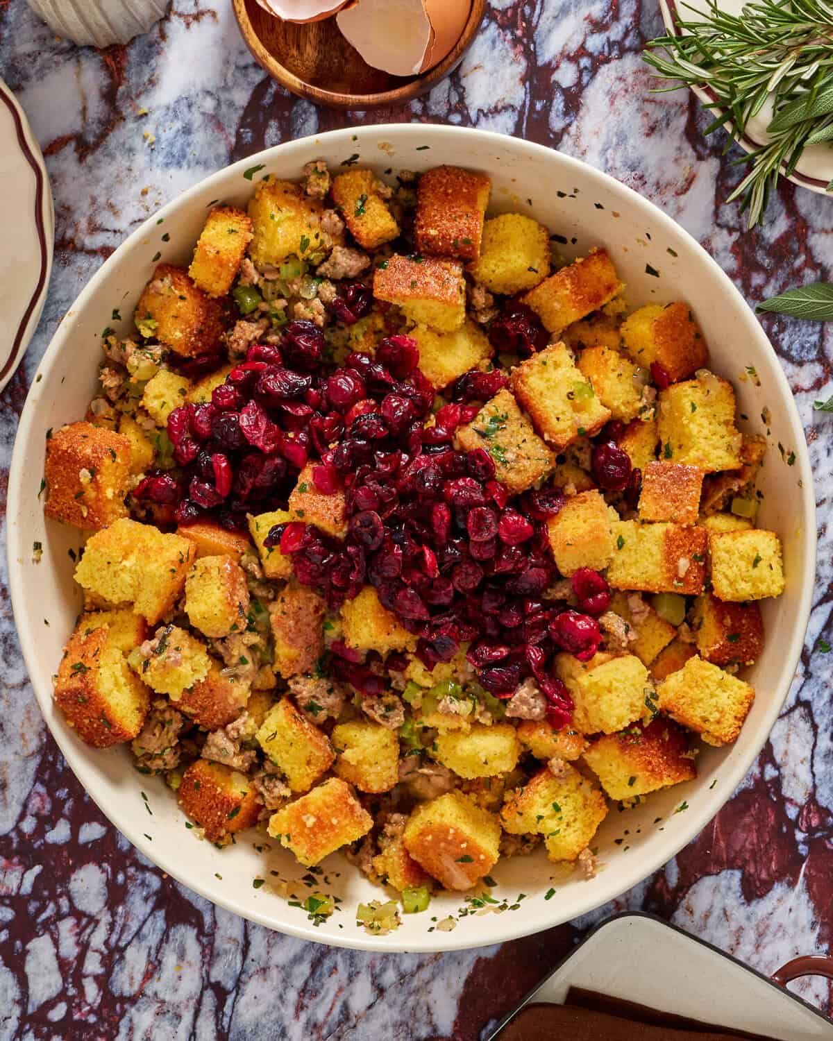 A large bowl filled with golden cornbread cubes, herbs, and crumbled sausage, topped with a generous pile of dried cranberries, sits on a marble countertop with rosemary and egg shells nearby.