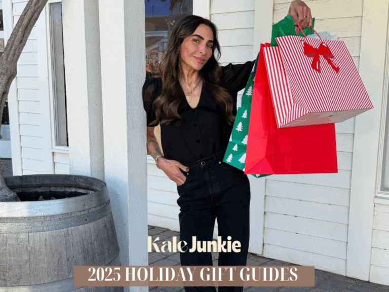 A woman in black clothing smiles while holding several colorful gift bags. Text on the image reads “Kale Junkie 2025 Holiday Gift Guides.” She stands outside a white building near a large planter.