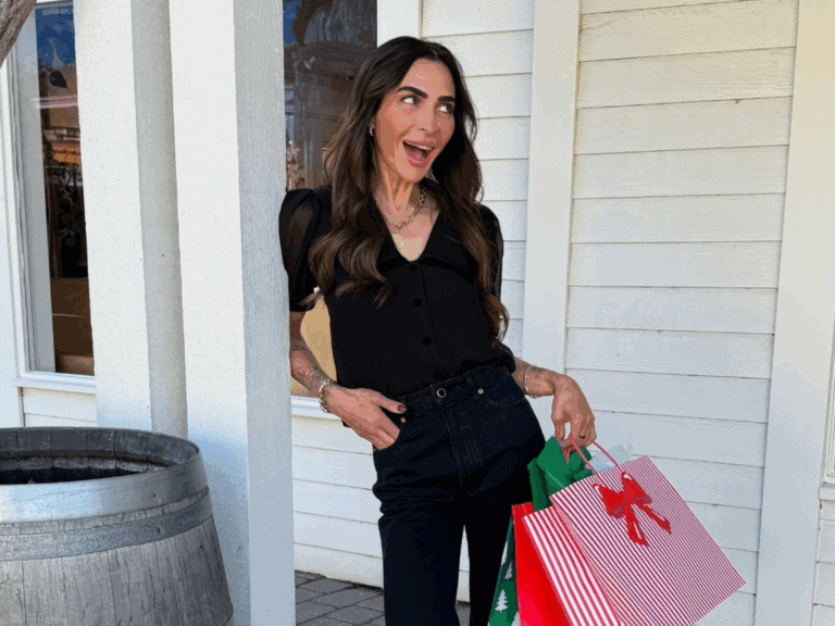 A smiling woman with long dark hair stands outside a white building, holding festive shopping bags. She is dressed in a black blouse and dark jeans, looking to the side with a joyful expression.