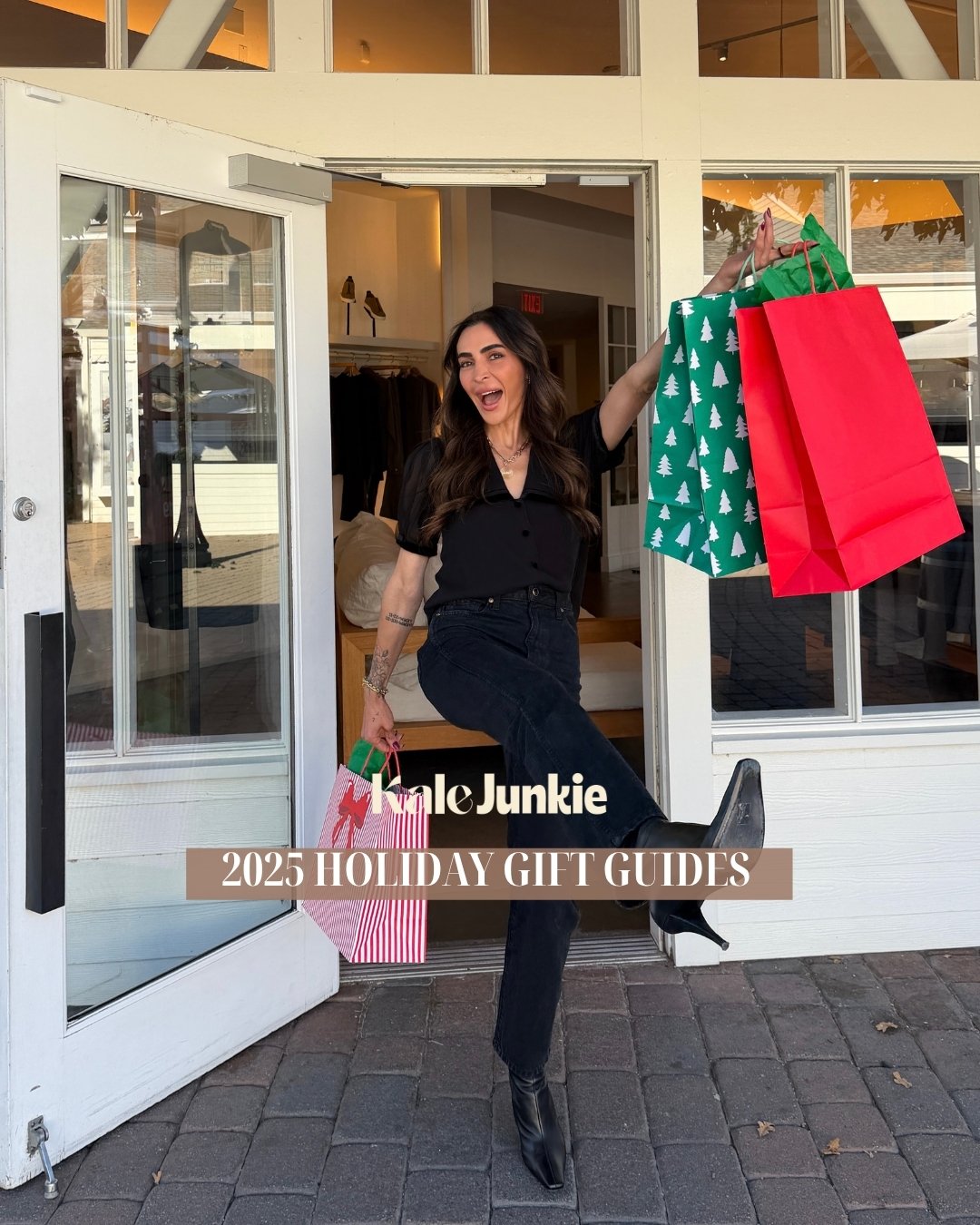 A smiling woman with long dark hair, dressed in black, holds several colorful holiday shopping bags and kicks up her boot in excitement outside a store. Text reads “2025 HOLIDAY GIFT GUIDES.”.