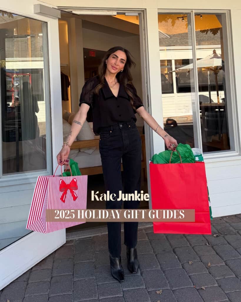 A woman in black clothes stands outside a store holding large holiday gift bags. Text on the image reads "Kale Junkie 2025 Holiday Gift Guides." The scene suggests holiday shopping.