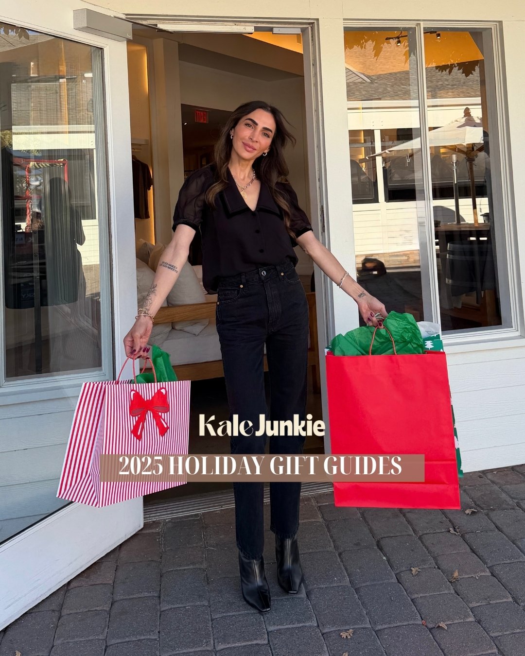 A woman in black clothes stands outside a store holding large holiday gift bags. Text on the image reads "Kale Junkie 2025 Holiday Gift Guides." The scene suggests holiday shopping.