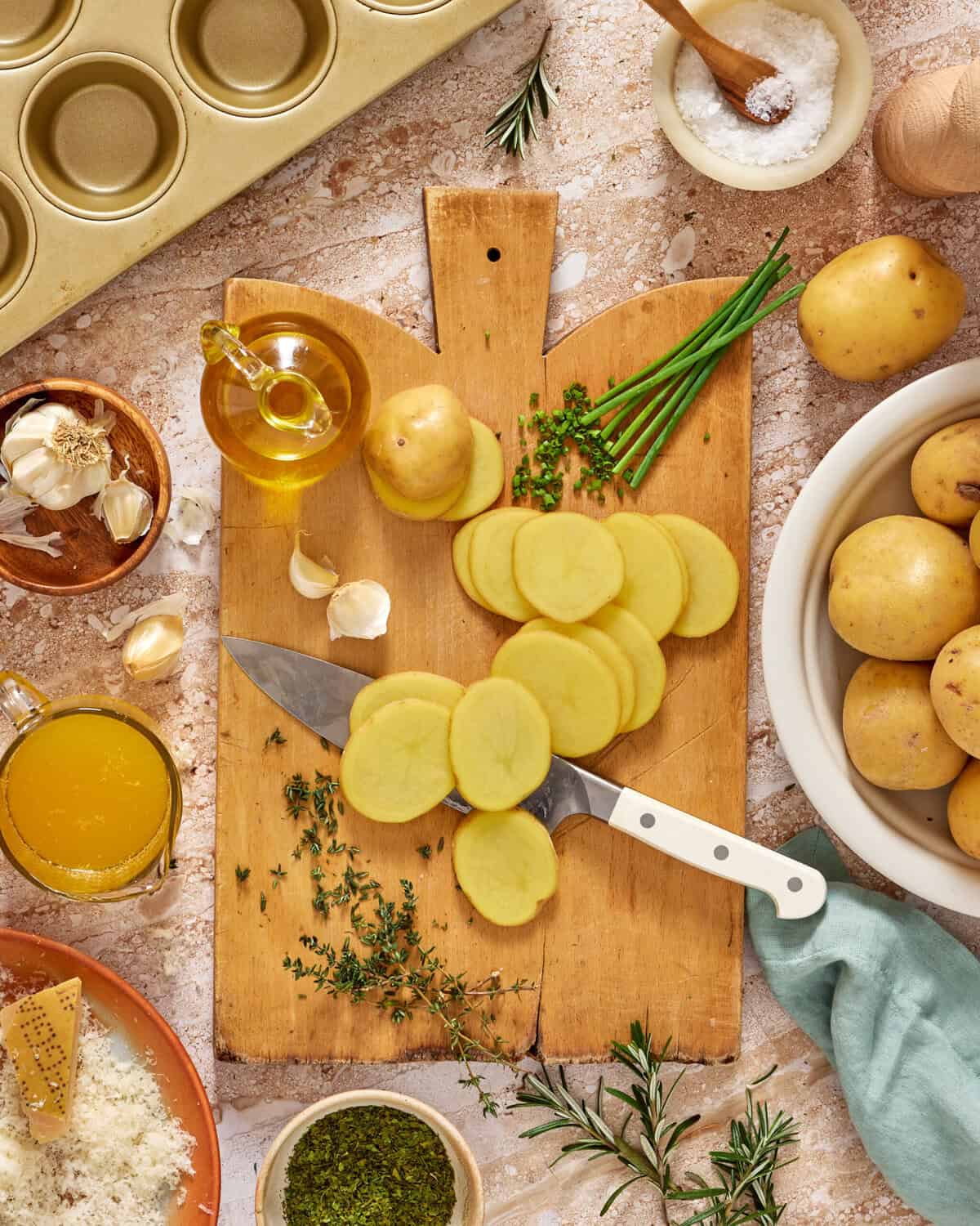 Sliced potatoes on a wooden cutting board with a knife, surrounded by whole potatoes, herbs, garlic, olive oil, parmesan, salt, and a muffin tin on a rustic kitchen surface.