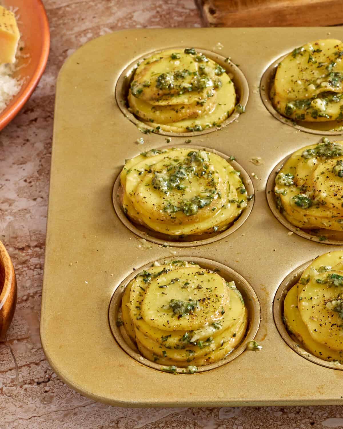 A muffin tin holds six stacks of thinly sliced potatoes topped with herbs and seasoning, ready to be baked. The background shows a rustic countertop with a bowl of grated cheese and some scattered seasonings.