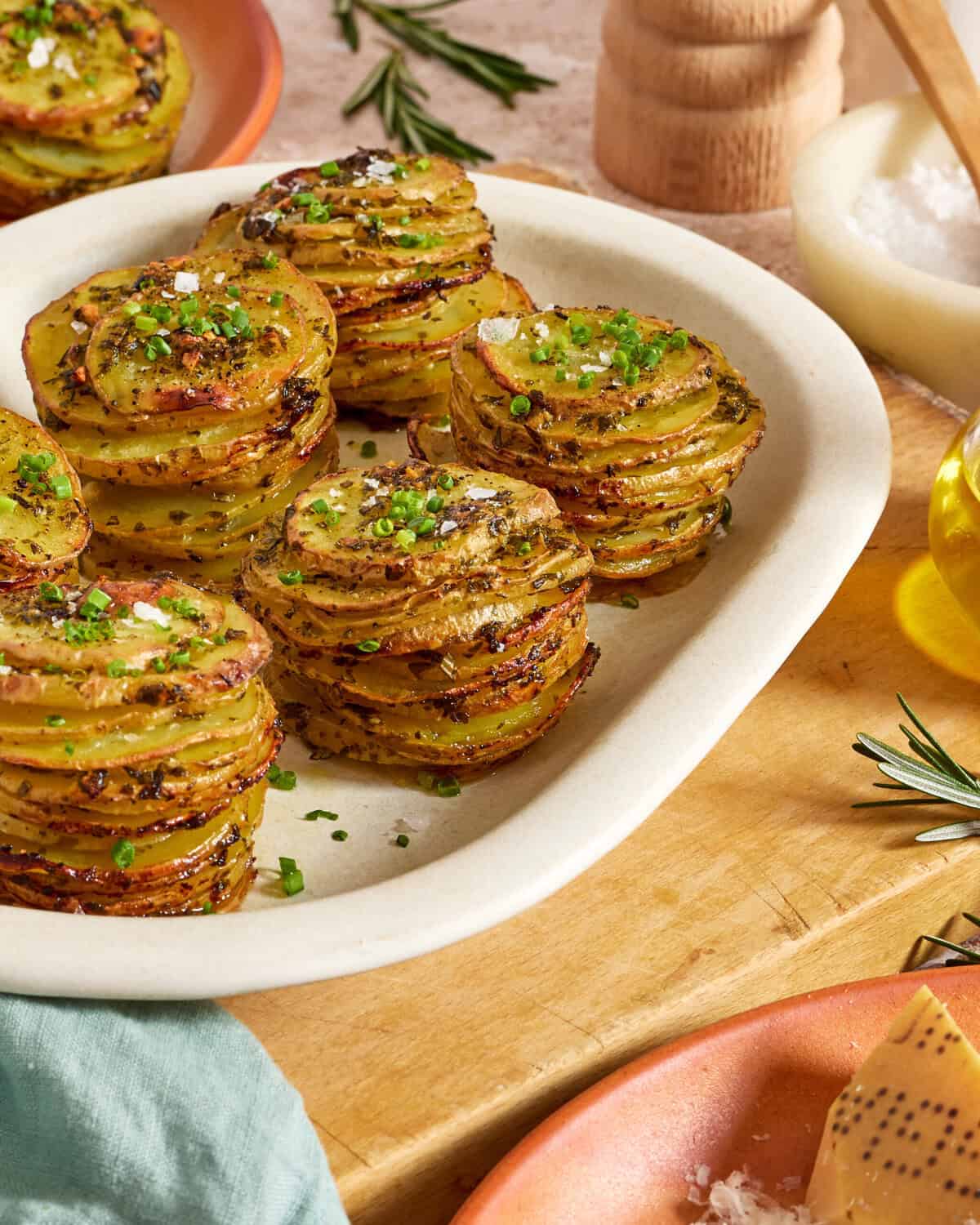 A plate of stacked, thinly sliced roasted potatoes garnished with chopped chives and sea salt. Fresh rosemary, olive oil, and a bowl of salt are nearby on a wooden surface.