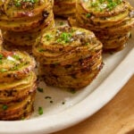 Stacks of thinly sliced, roasted potatoes garnished with chopped herbs and coarse salt are arranged neatly on an off-white oval serving dish.