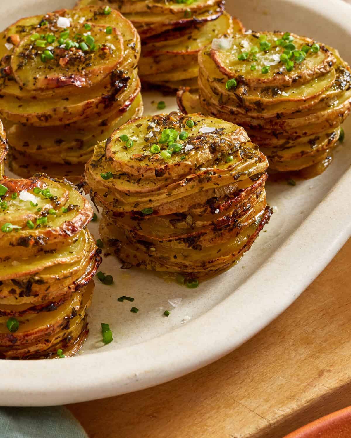 Stacks of thinly sliced, roasted potatoes garnished with chopped herbs and coarse salt are arranged neatly on an off-white oval serving dish.