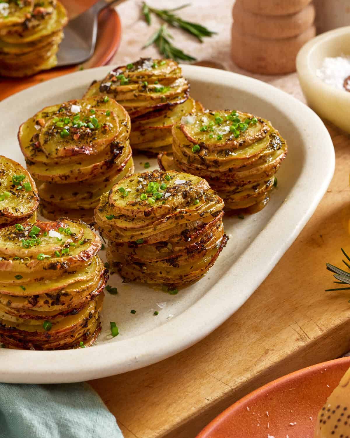 A white plate with stacks of thinly sliced roasted potatoes, seasoned with herbs and garnished with chopped chives, sits on a wooden surface with rosemary, salt, and pepper in the background.