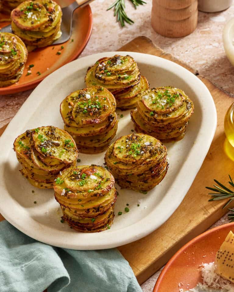 A plate of stacked, thinly sliced roasted potatoes garnished with chopped herbs sits on a table, surrounded by a green napkin, olive oil, fresh rosemary, and plates.