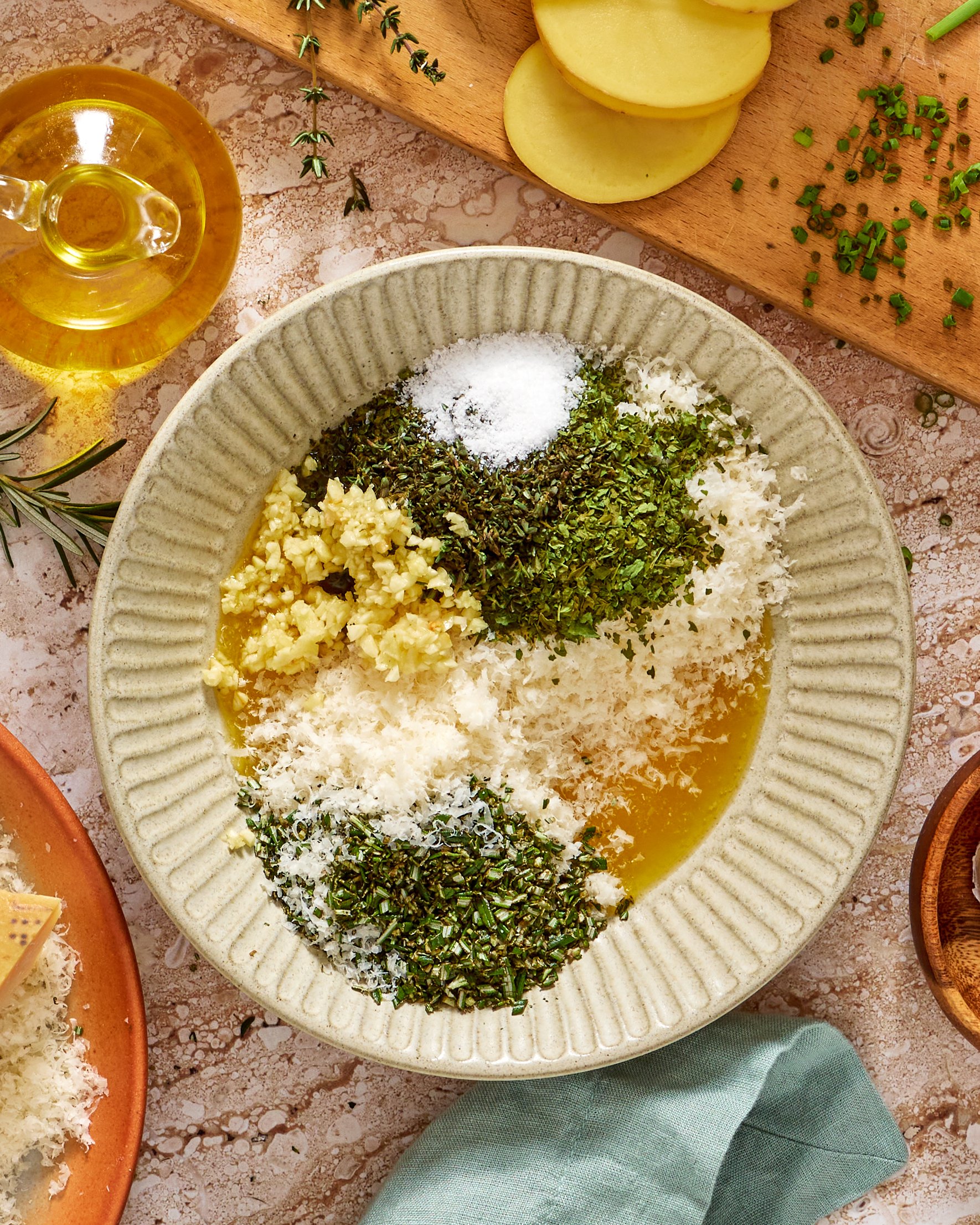 A bowl contains grated cheese, chopped herbs, minced garlic, salt, and oil. Surrounding the bowl are fresh herbs, a wooden board with sliced potatoes and chives, a small glass jug of oil, and a blue cloth.