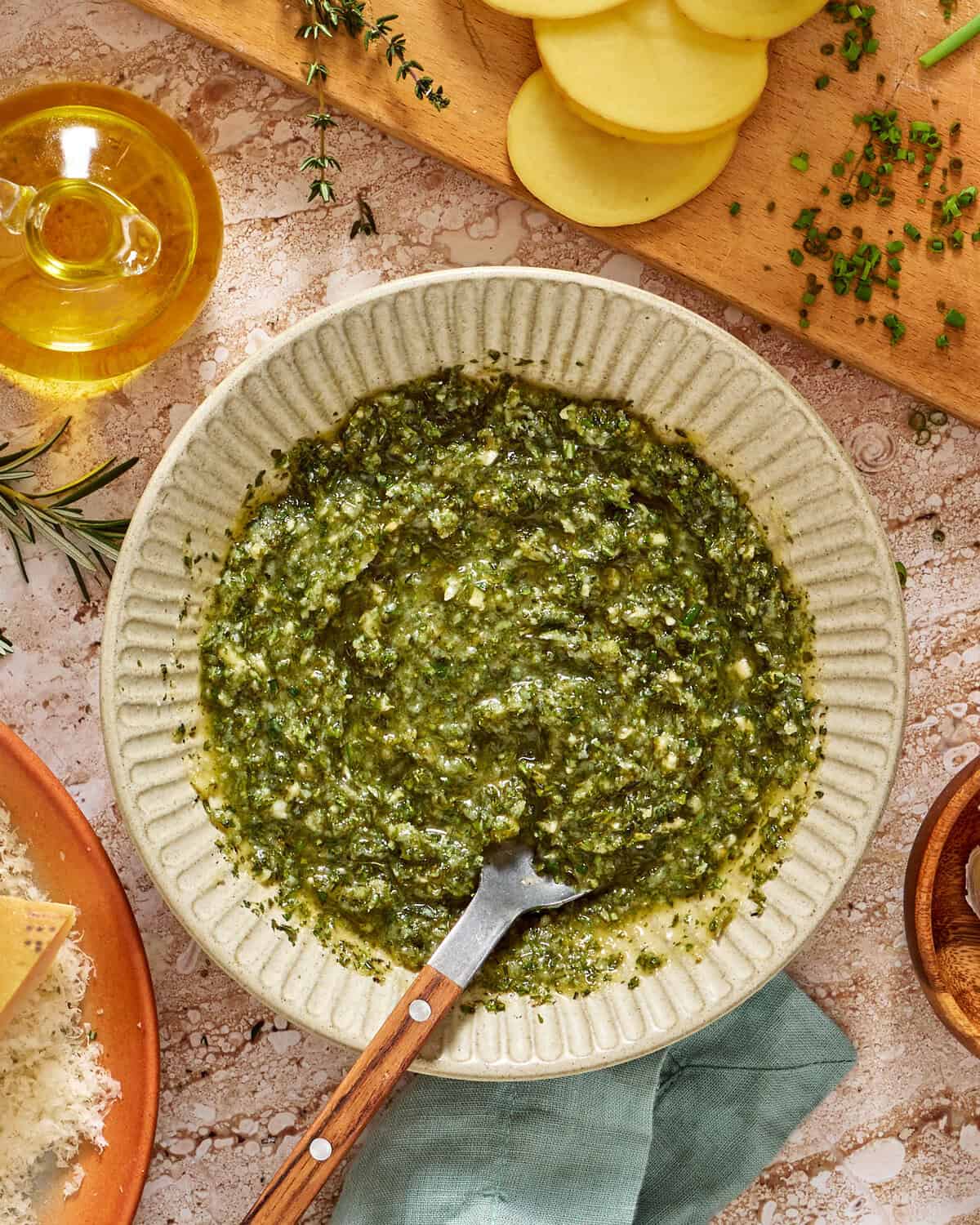 A bowl of green pesto sauce with a spoon, surrounded by olive oil, sliced potatoes, fresh herbs, grated cheese, and a wooden cutting board on a rustic surface.