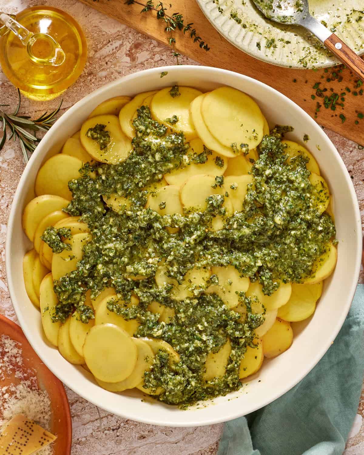 A white dish filled with thinly sliced potatoes topped with green pesto sauce, surrounded by a bottle of olive oil, fresh herbs, grated cheese, and a spoon on a rustic surface.