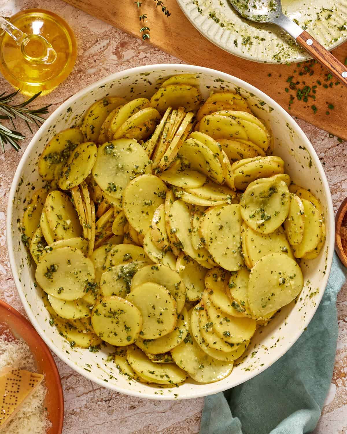 A large white bowl filled with thinly sliced potatoes coated in green herb seasoning, surrounded by ingredients like olive oil, parmesan, and fresh herbs on a light-colored table.