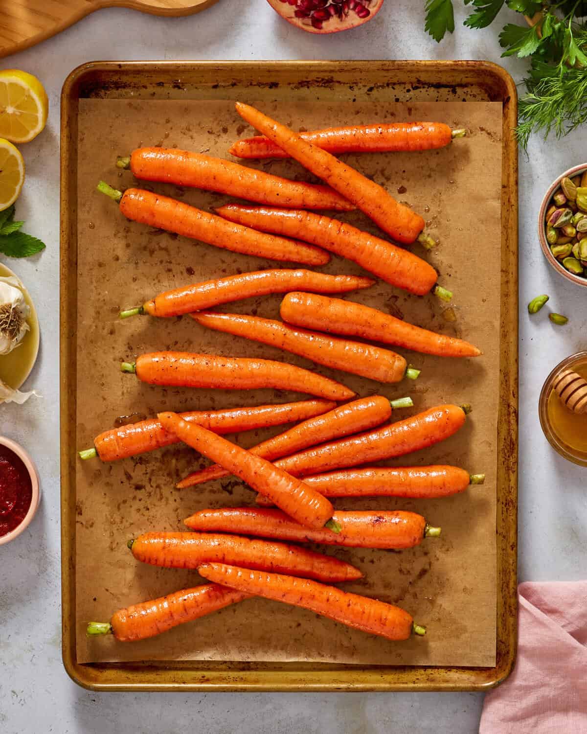 A baking sheet lined with parchment paper holds several whole, unpeeled carrots arranged in rows. Surrounding the tray are ingredients like lemon, parsley, garlic, pistachios, and a bowl of honey.