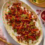 An oval platter with roasted carrots, crispy chickpeas, green herb sauce, and pomegranate seeds on a bed of creamy dip, garnished with herbs. Other small bowls with sauce and pomegranate seeds are nearby.