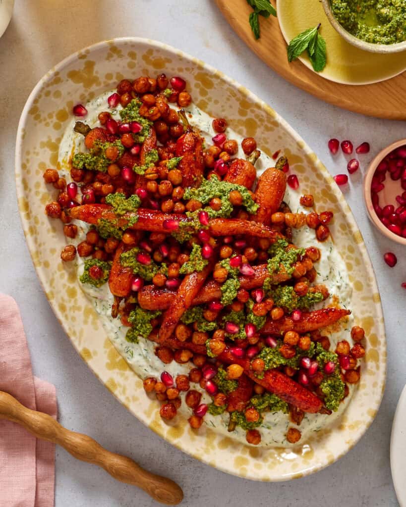 An oval platter with roasted carrots, crispy chickpeas, green herb sauce, and pomegranate seeds on a bed of creamy dip, garnished with herbs. Other small bowls with sauce and pomegranate seeds are nearby.