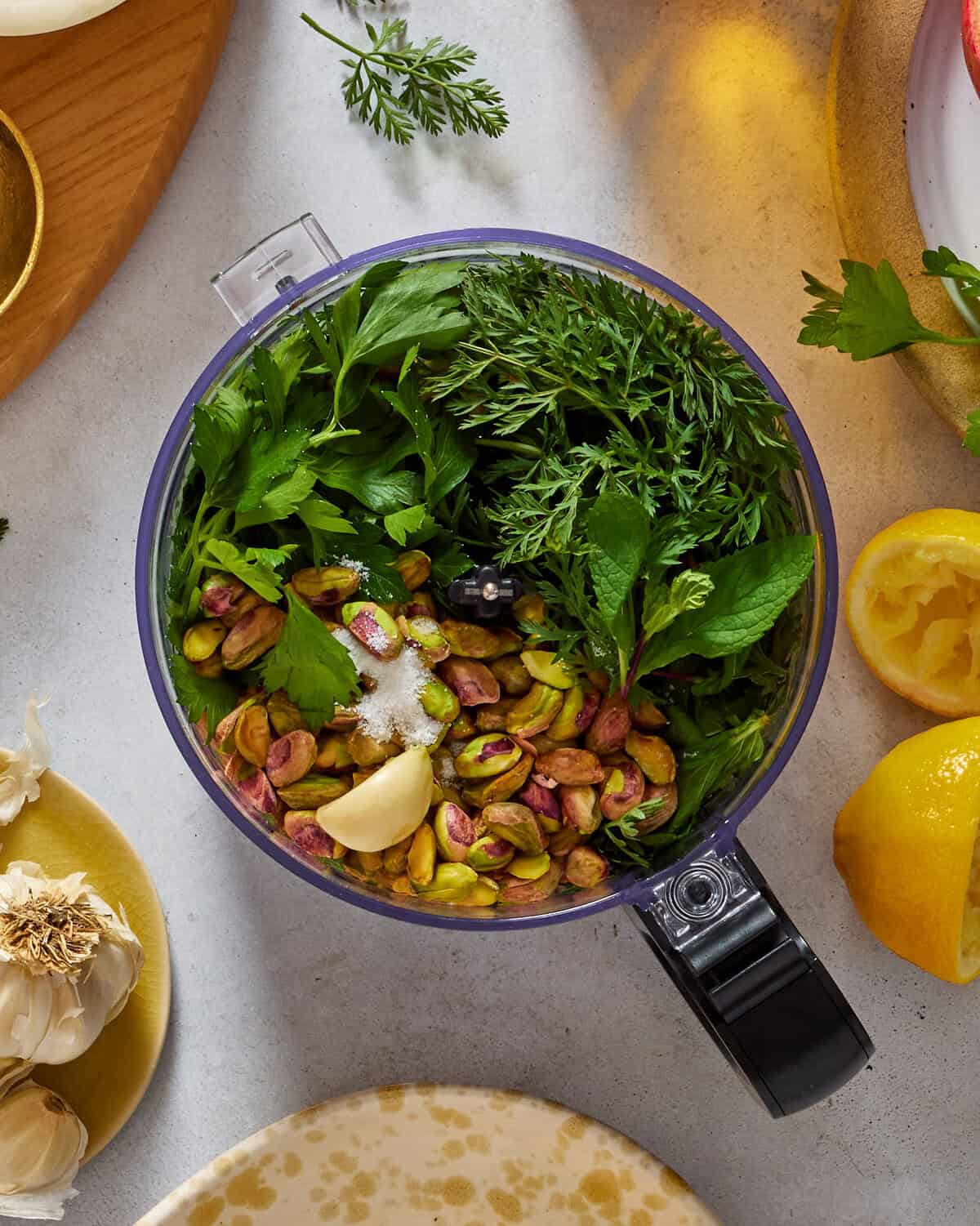 A food processor bowl filled with fresh herbs, pistachios, salt, and garlic cloves sits on a countertop surrounded by lemons, garlic bulbs, a plate, and a wooden board with parsley.