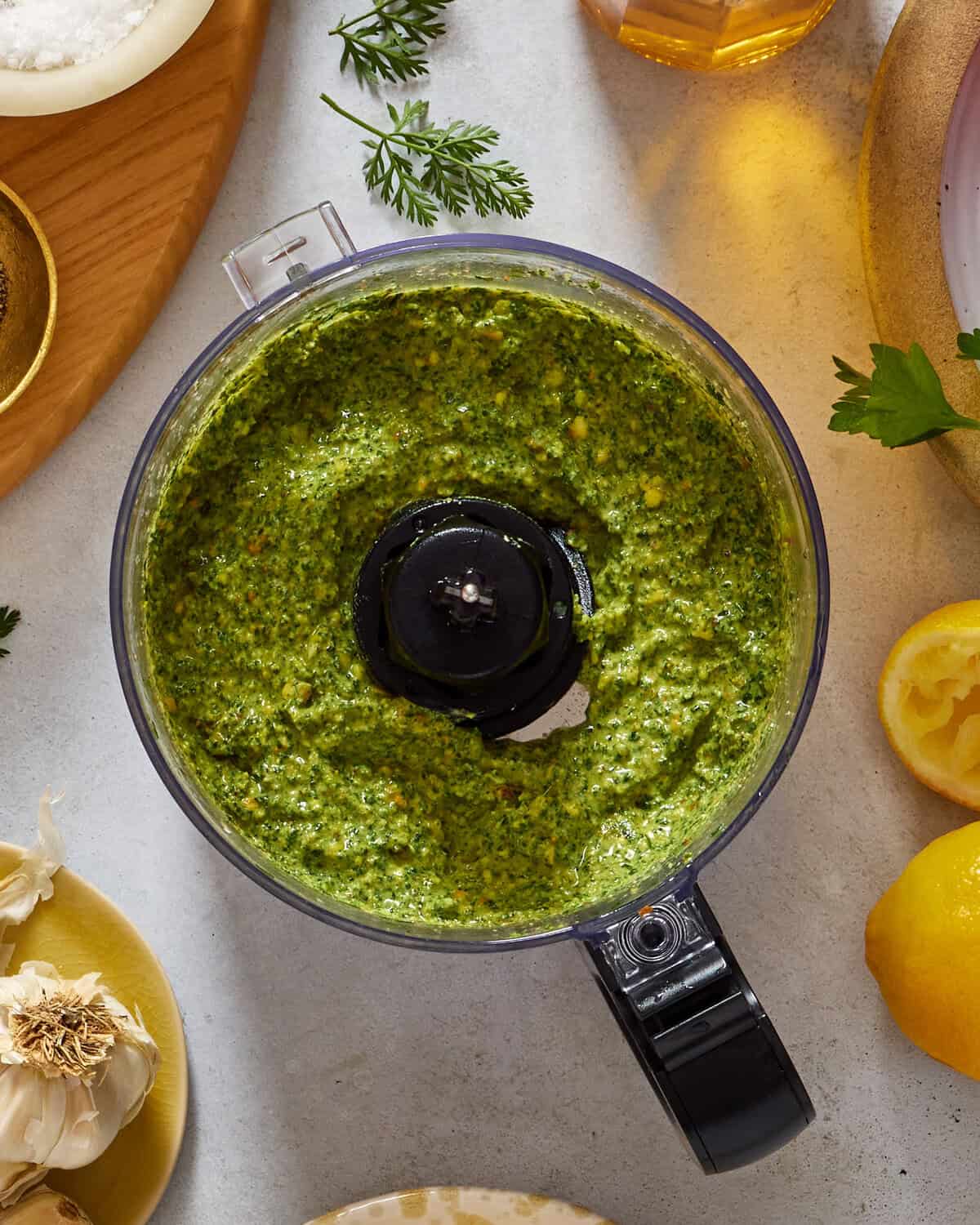 A food processor filled with freshly made green pesto sits on a countertop surrounded by garlic, lemon, parsley, and other ingredients.