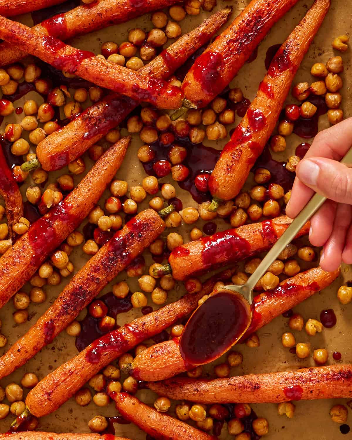 Roasted carrots and chickpeas on a baking tray, with a hand drizzling dark red sauce over the carrots using a spoon.