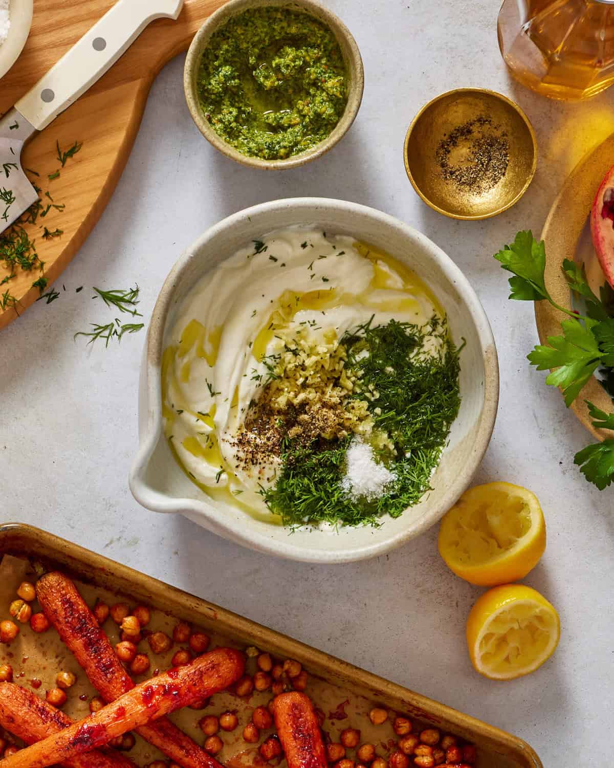 A bowl with yogurt, chopped herbs, minced garlic, black pepper, salt, and olive oil on a counter. Nearby are halved lemons, roasted carrots with chickpeas, pesto, a gold bowl with pepper, and fresh parsley.