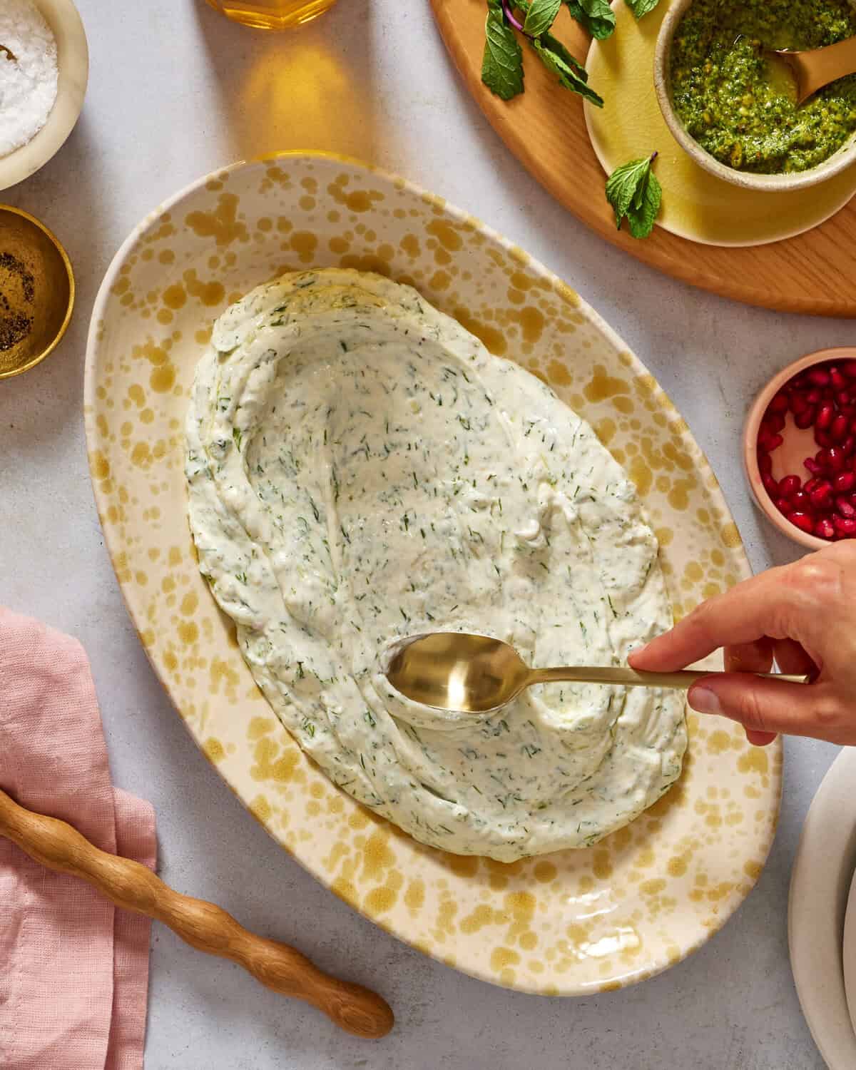 A hand uses a spoon to spread herbed yogurt on a large oval platter. Surrounding the platter are small bowls with salt, pepper, pesto, and pomegranate seeds, along with a pink napkin and a wooden rolling pin.