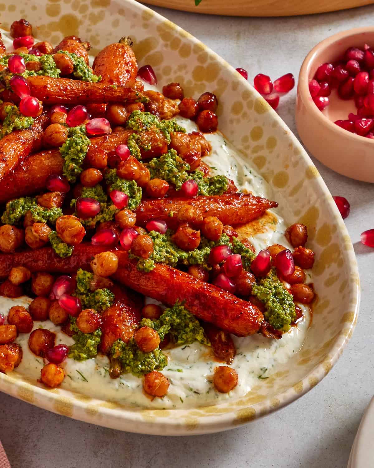 A dish of roasted carrots and chickpeas topped with green pesto and pomegranate seeds, served over a creamy white sauce with herbs, in an oval patterned bowl. A small bowl of pomegranate seeds sits nearby.