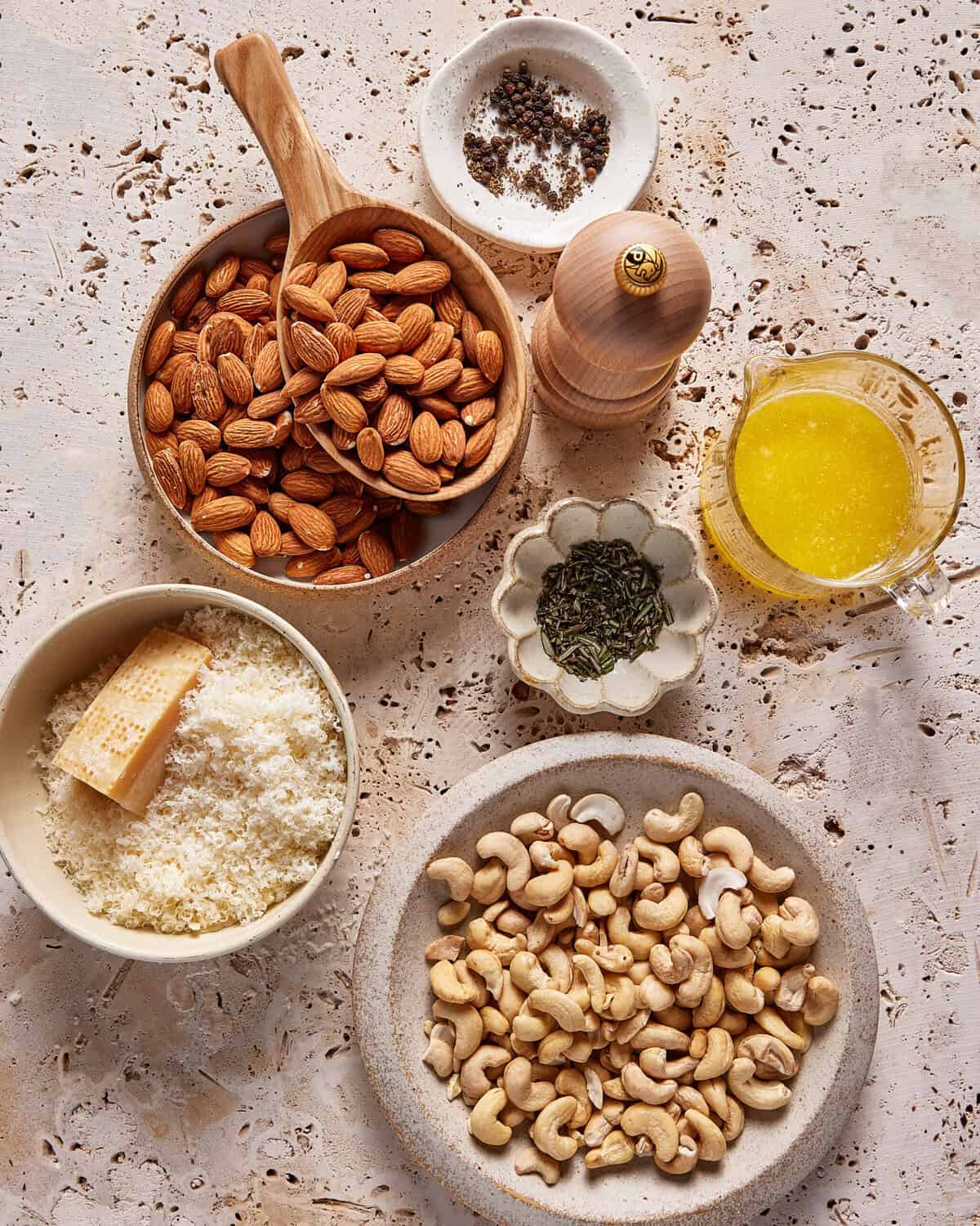 Assorted ingredients on a textured surface: a bowl of whole almonds with a wooden scoop, a bowl of cashews, grated and block Parmesan cheese, melted butter in a glass cup, ground pepper and salt, and dried herbs.