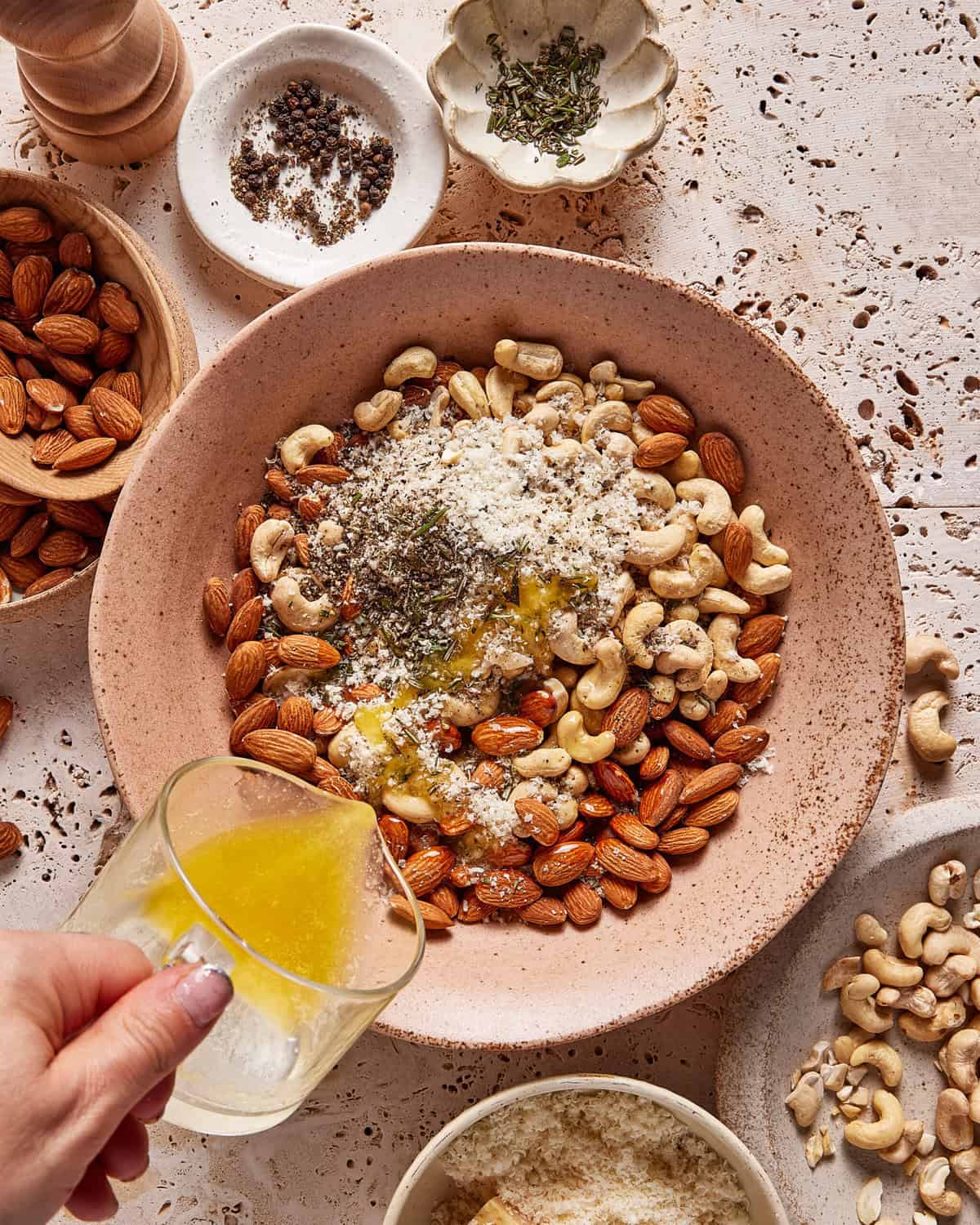 A hand pours melted butter from a glass cup onto a bowl filled with raw cashews, almonds, grated cheese, and herbs. Various small bowls of nuts, spices, and cheese surround the main bowl on a speckled countertop.