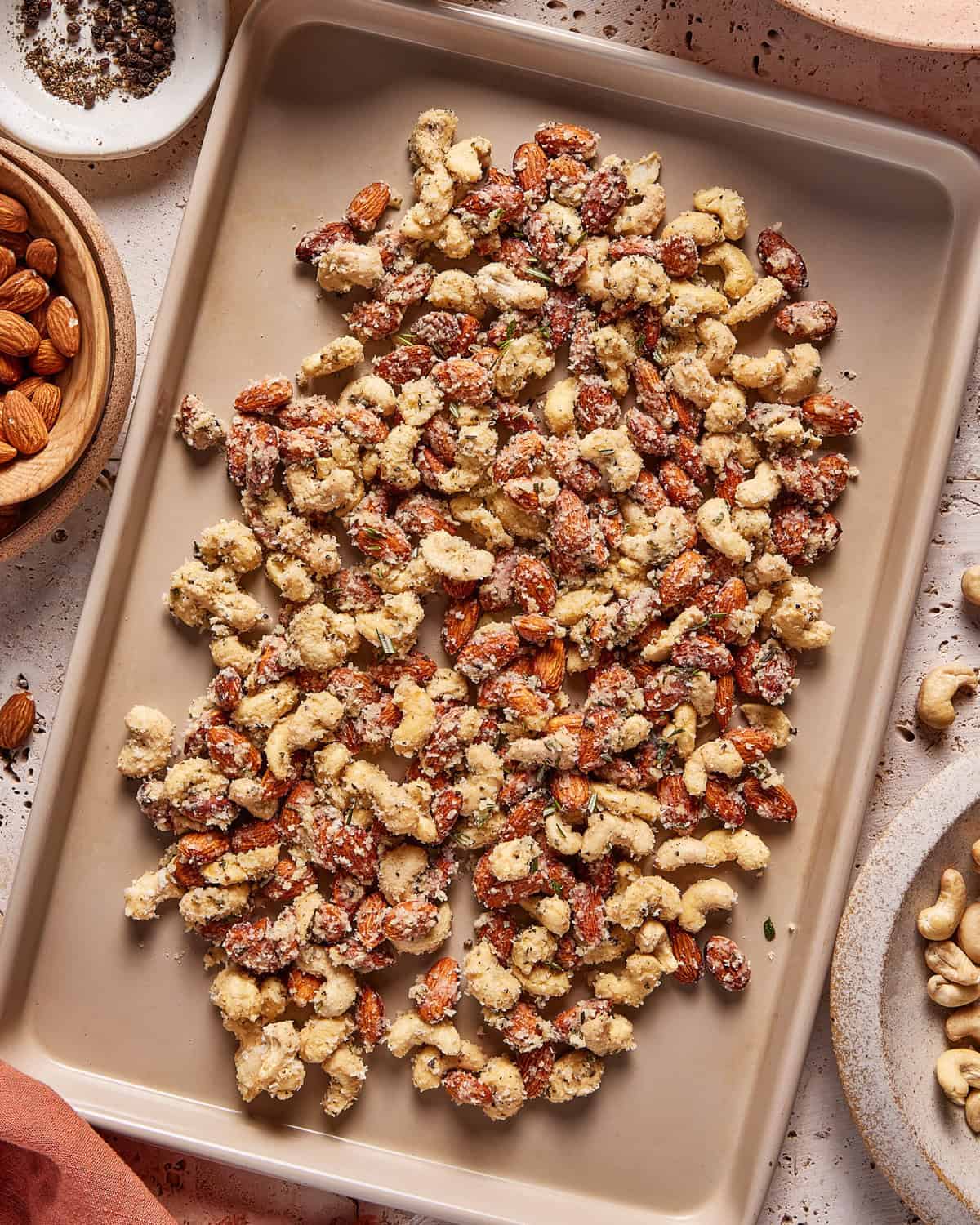 A baking tray filled with a mix of roasted nuts coated in a light seasoning. Bowls of cashews, almonds, and spices are visible nearby on a beige surface.