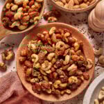 A plate and wooden spoon filled with mixed roasted nuts, including almonds and cashews, garnished with herbs. Surrounding the plate are bowls of plain almonds and more mixed nuts on a decorative surface.
