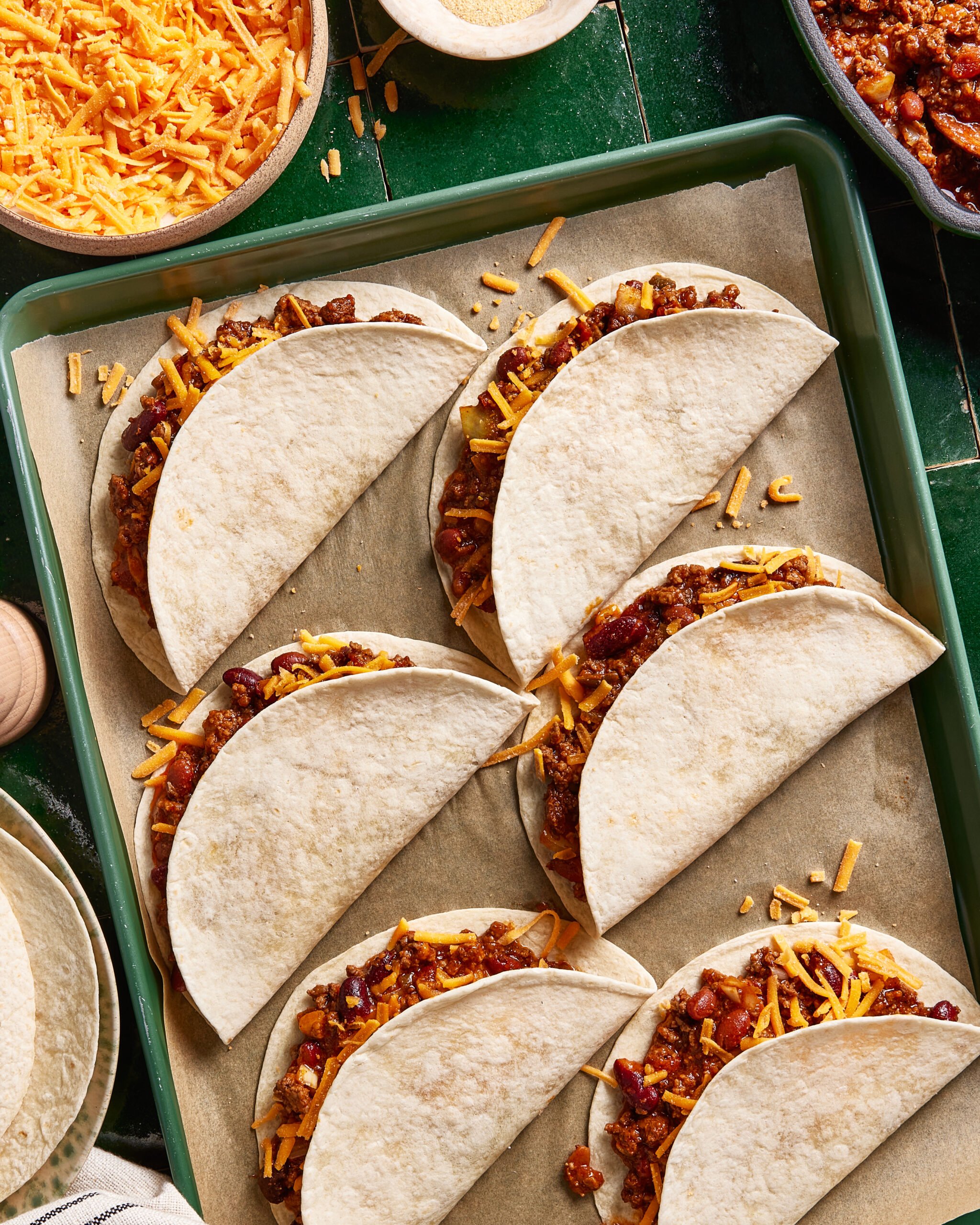 Six soft tacos filled with ground meat chili and topped with shredded cheddar cheese are arranged on a parchment-lined baking sheet. Bowls of extra cheese and chili are nearby.