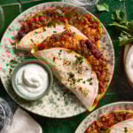 A plate with two folded tortillas filled with chili and melted cheese, garnished with chopped parsley, next to a small bowl of sour cream, on a green-tiled table.