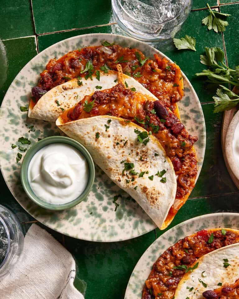 A plate with two folded tortillas filled with chili and melted cheese, garnished with chopped parsley, next to a small bowl of sour cream, on a green-tiled table.