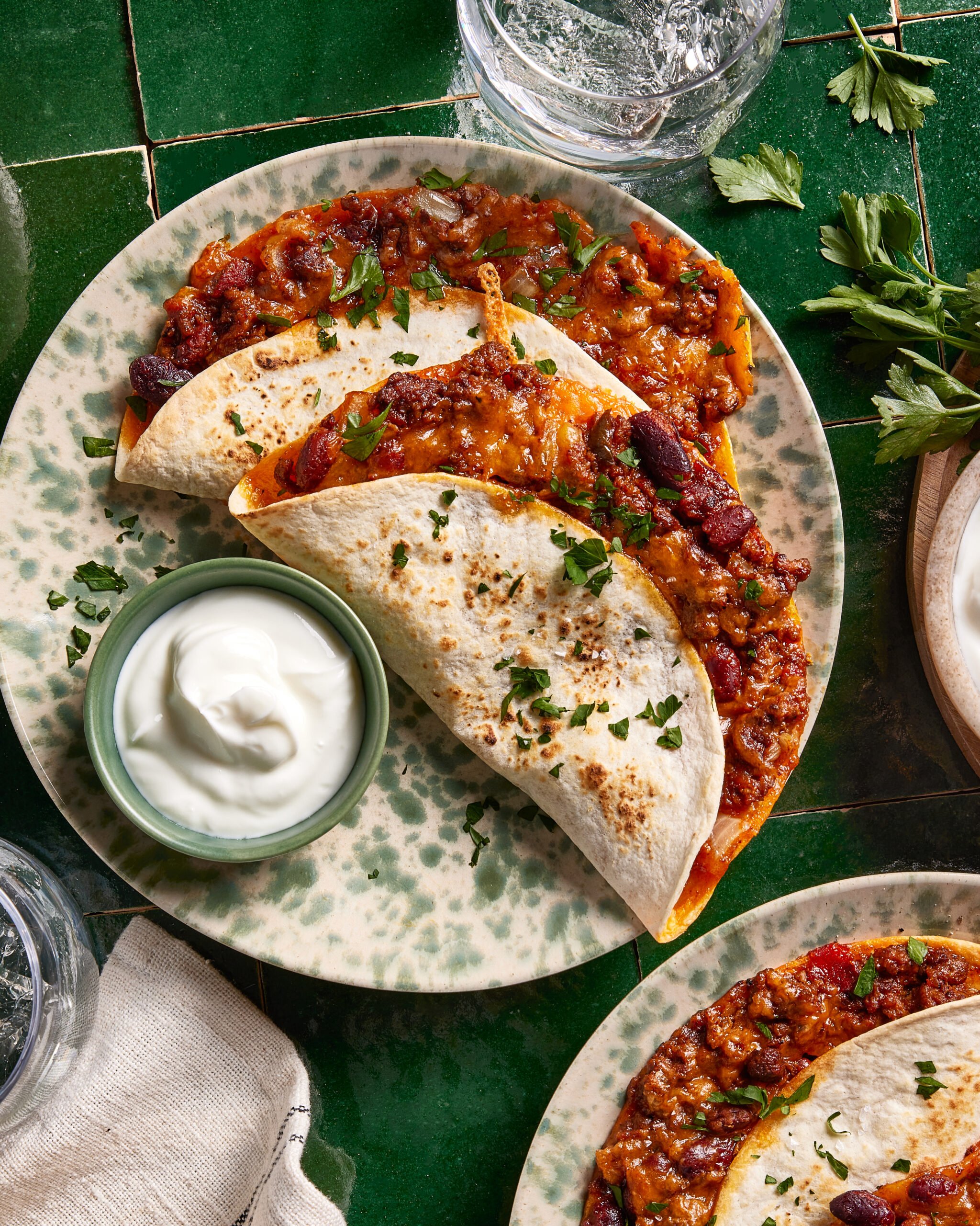 A plate with two folded tortillas filled with chili and melted cheese, garnished with chopped parsley, next to a small bowl of sour cream, on a green-tiled table.