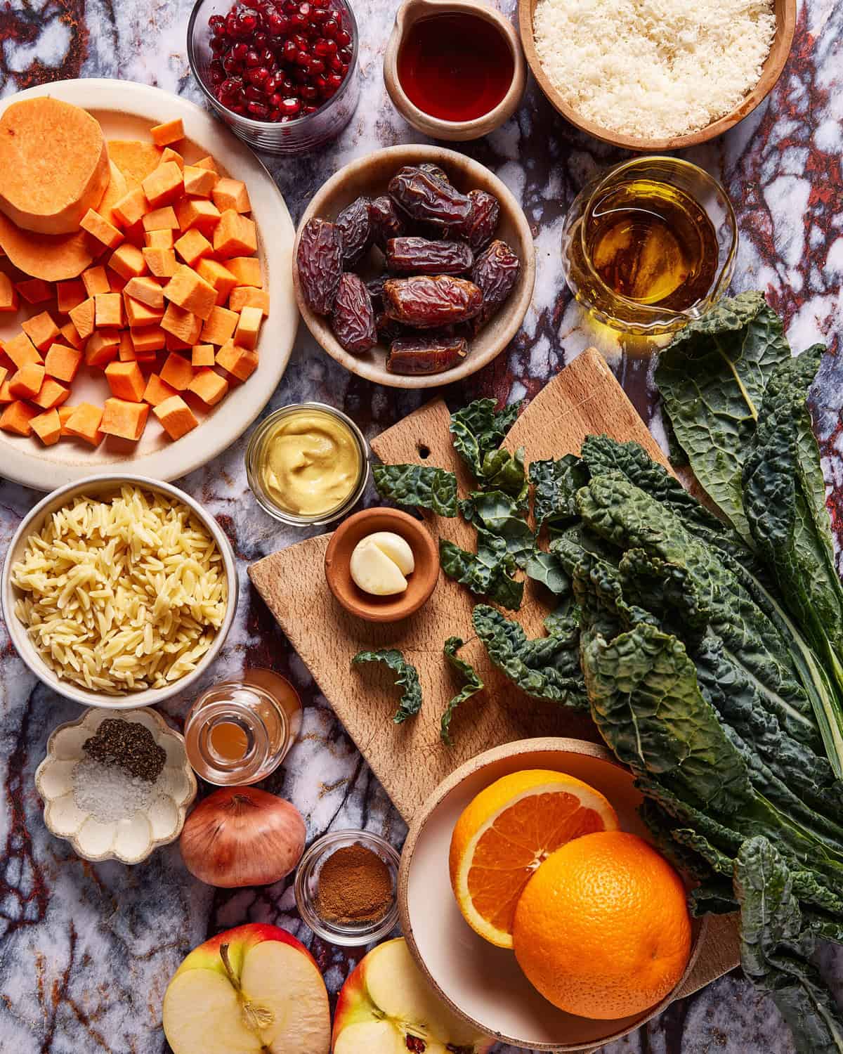 Overhead view of assorted ingredients on a marble surface, including chopped sweet potatoes, kale, orzo pasta, dates, sliced apple, orange halves, pomegranate seeds, garlic, olive oil, spices, and mustard.