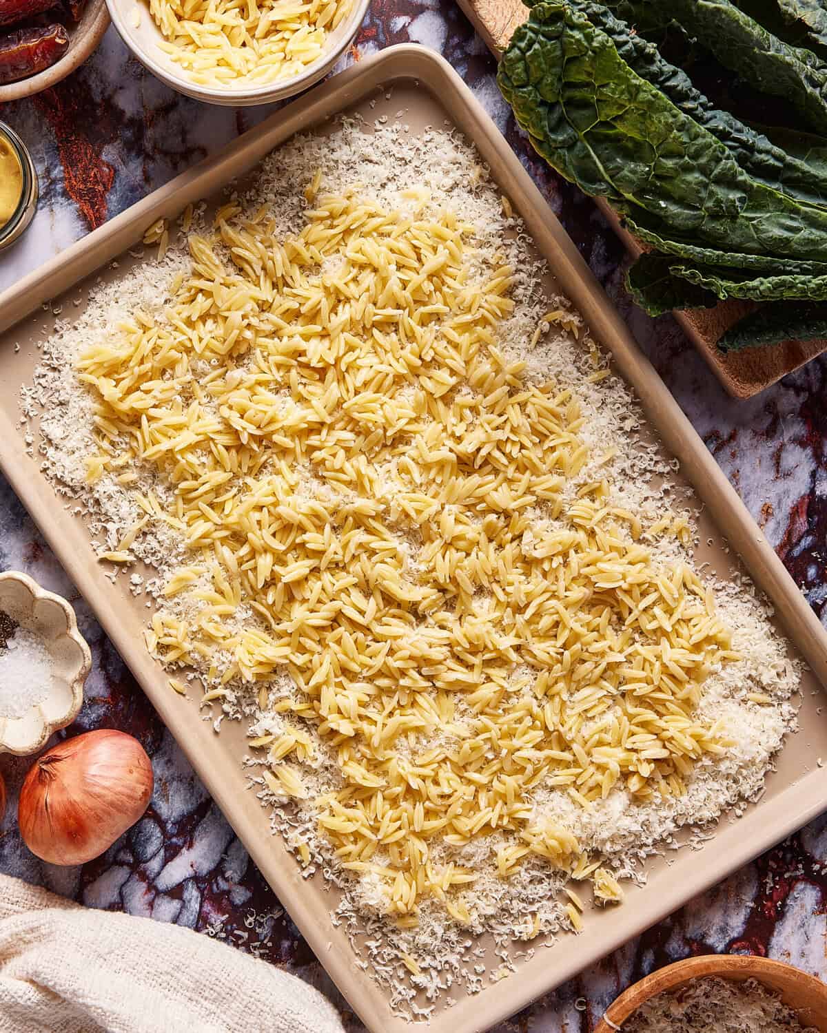 A baking sheet filled with uncooked orzo pasta and grated cheese, surrounded by ingredients like kale leaves, a shallot, a bowl of oil, salt, and a bowl of grated cheese on a marble countertop.