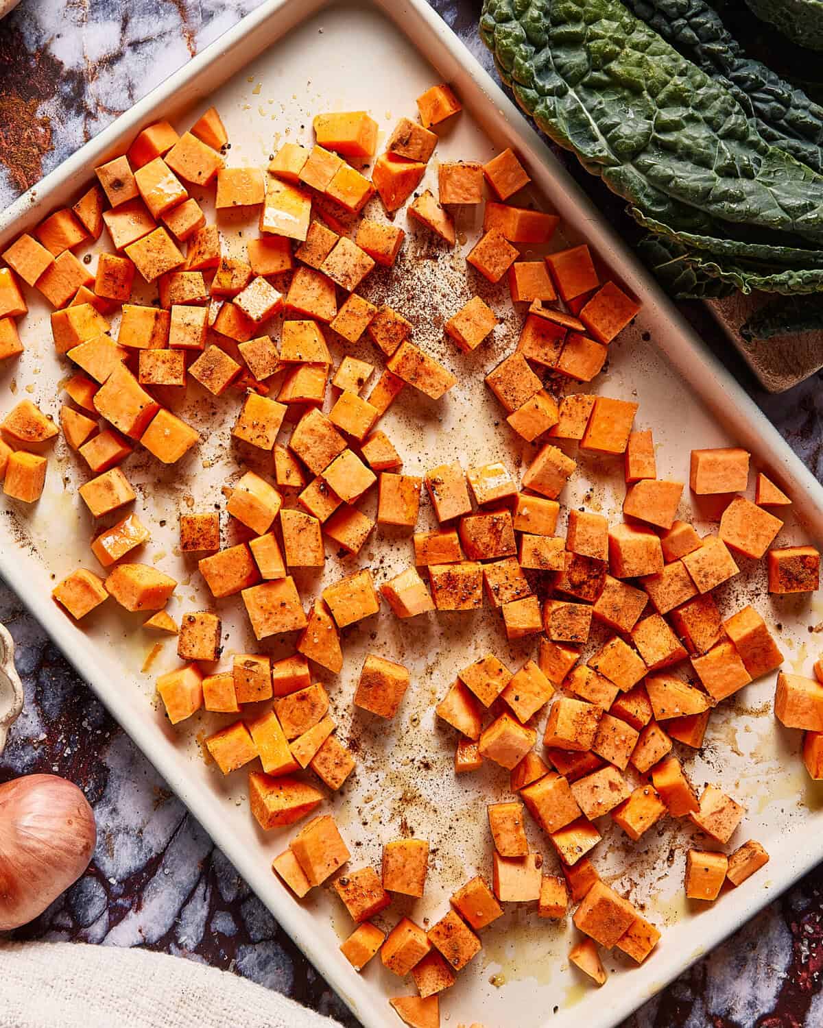 A baking sheet filled with evenly spread, cubed sweet potatoes seasoned with spices; leafy greens, a shallot, and garlic are visible on the side.
