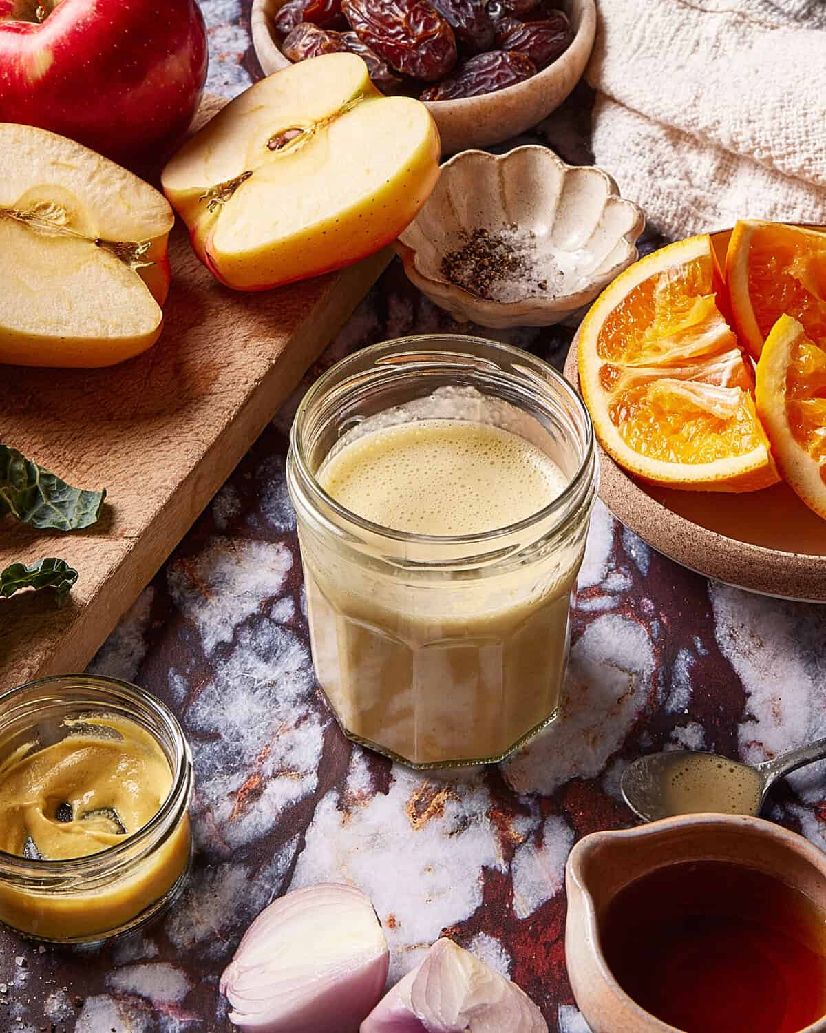 A jar of creamy dressing sits on a marble surface surrounded by sliced apples, orange wedges, a small bowl of mustard, shallot, a bowl of dates, and a small dish of pepper.