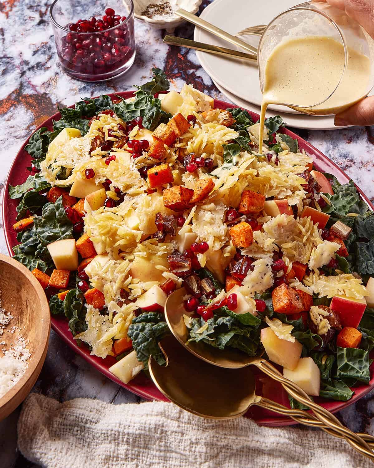 A colorful salad with kale, shredded cabbage, roasted sweet potatoes, apple chunks, pomegranate seeds, and nuts on a red platter. A hand pours dressing over the salad; spoons, salt, and pomegranate seeds are nearby.