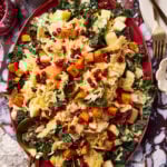 A colorful salad on a red platter with kale, roasted butternut squash cubes, diced apples, shredded cabbage, pomegranate seeds, and a creamy dressing, surrounded by cutlery and a glass of pink beverage on a marbled surface.