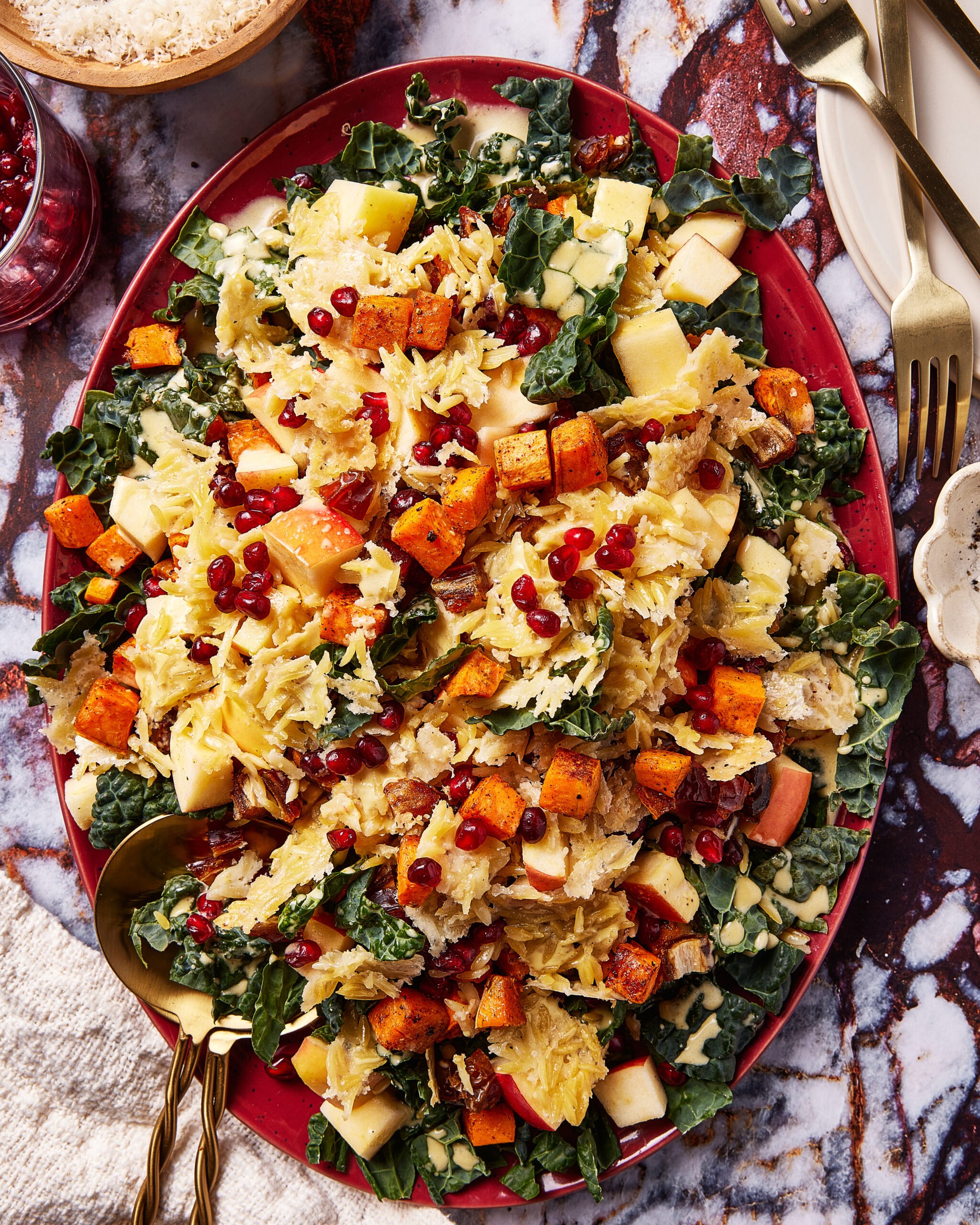 A colorful salad on a red platter with kale, roasted butternut squash cubes, diced apples, shredded cabbage, pomegranate seeds, and a creamy dressing, surrounded by cutlery and a glass of pink beverage on a marbled surface.