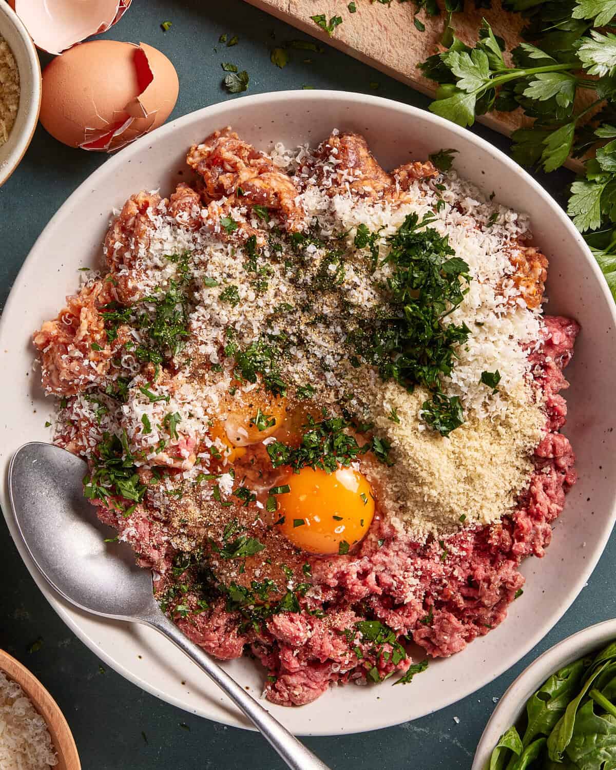 A large bowl contains ground meat, two raw eggs, grated cheese, breadcrumbs, chopped parsley, and seasonings, with a spoon resting on the side. Surrounding the bowl are eggshells and fresh parsley.